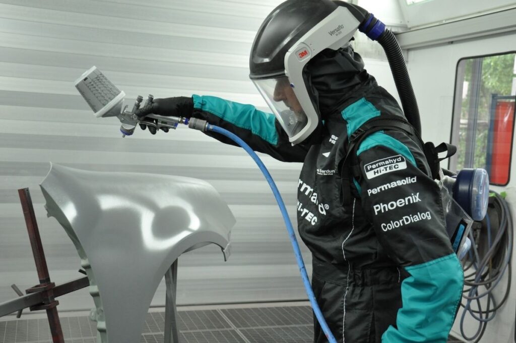 Covek u odelu za farbanje automobila koji uz pomoc pistolja farba krilo automobila